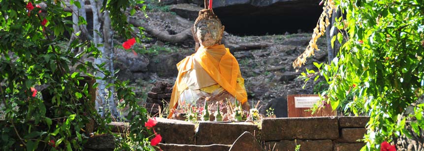 Sitzender Buddha aus Stein geschmückt mit gelbem Tuch und Opfergaben wie Räucherstäbchen im Vordergrund in der Tempelanlage von Wat Phou in Laos